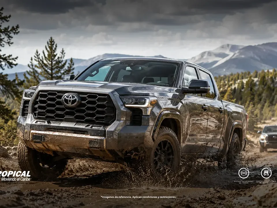 Mud-splattered gray Toyota pickup driving on a rocky dirt trail with mountains in the background. Branding: AUTOTROPICAL and Toyota.