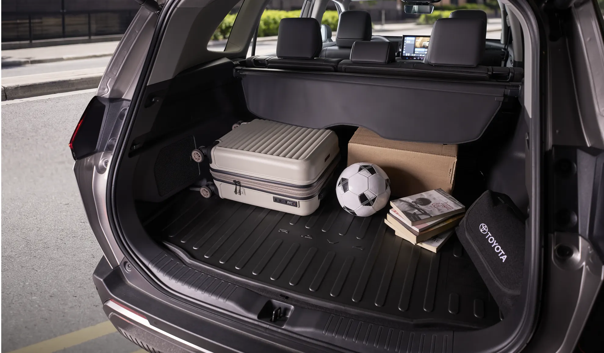 Open trunk of SUV with a beige rolling suitcase, a cardboard box, a soccer ball, and magazines visible in the cargo area.