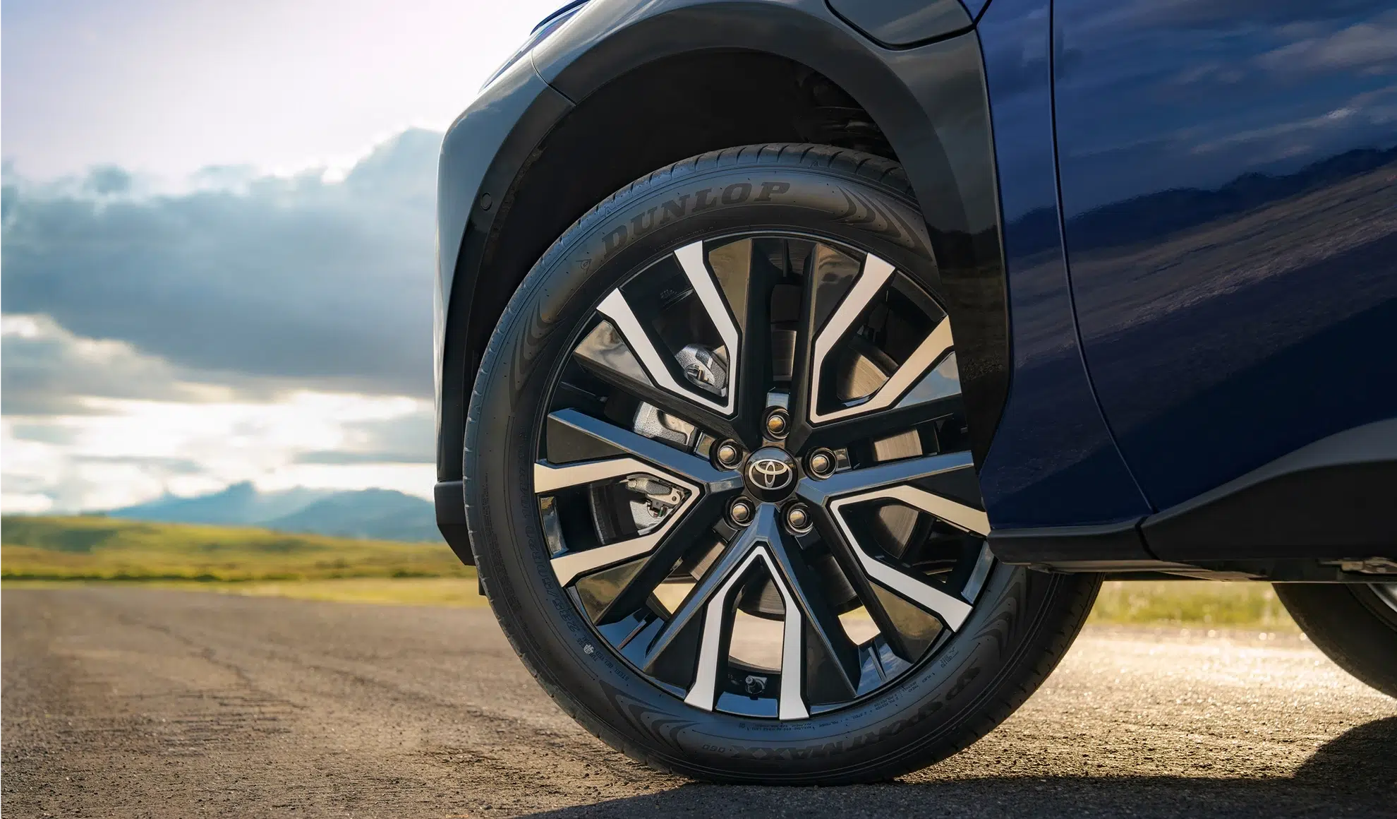 Close-up of a car wheel and front fender, showing a stylish alloy rim and Dunlop tire with a Toyota center cap on a blue vehicle under a cloudy sky.