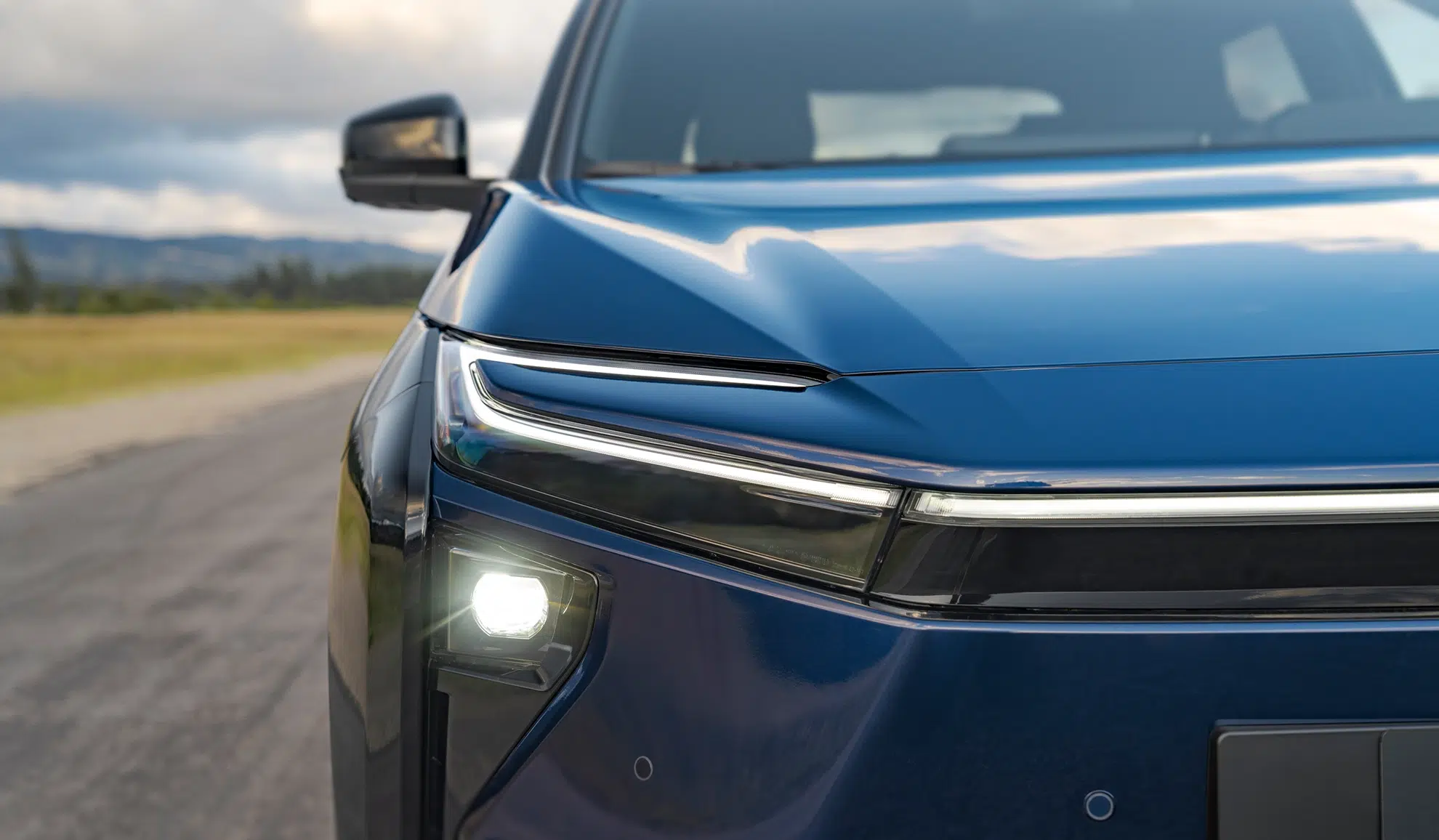 Close-up of the front left corner of a blue SUV, showing the headlight, glossy hood, and side mirror on a rural road.
