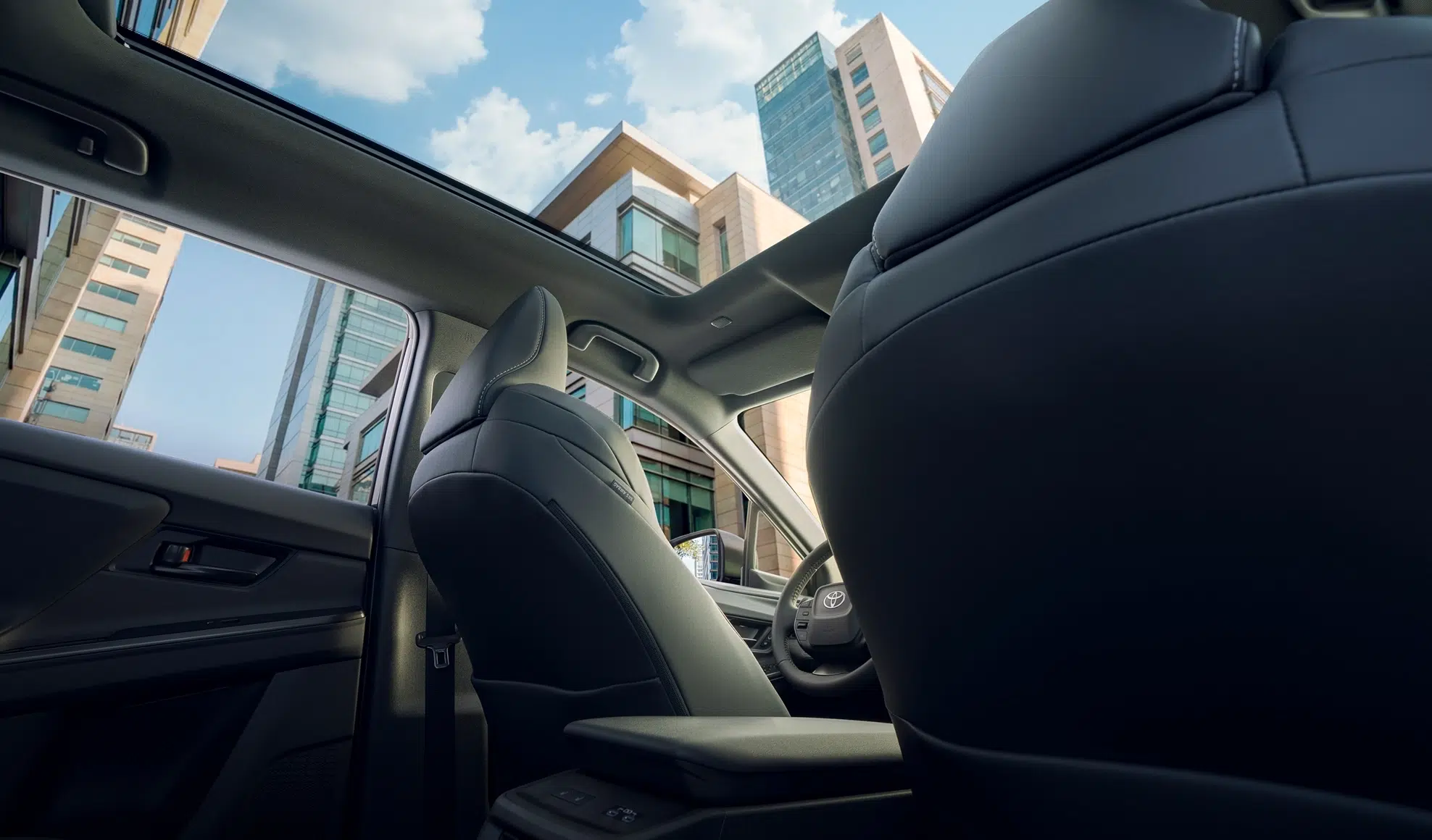 Rear-seat view of a car interior with dark leather seats and a panoramic sunroof, city buildings and blue sky above side windows.