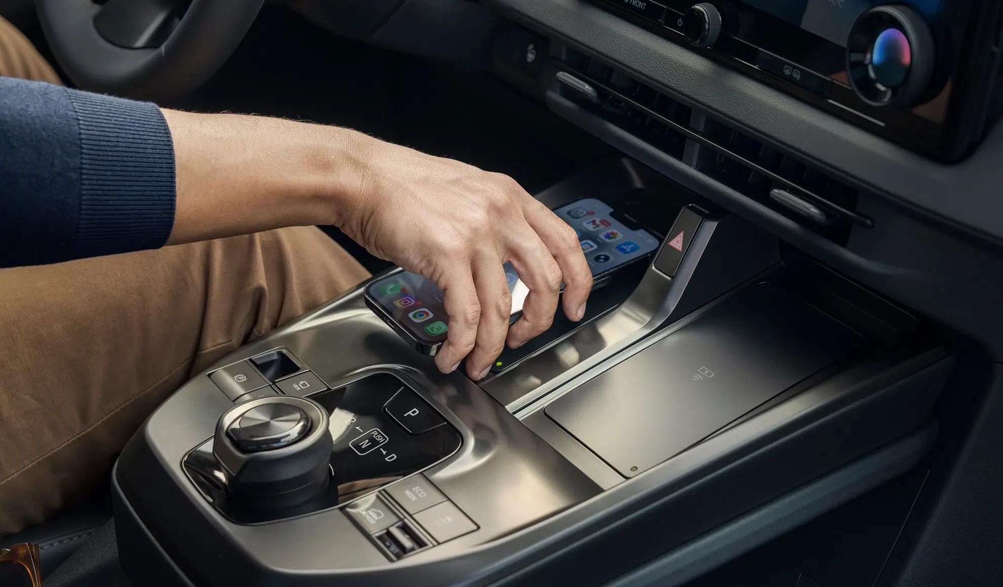 Person resting a smartphone on the car's center console wireless charging pad beside the gear shifter and dashboard controls.