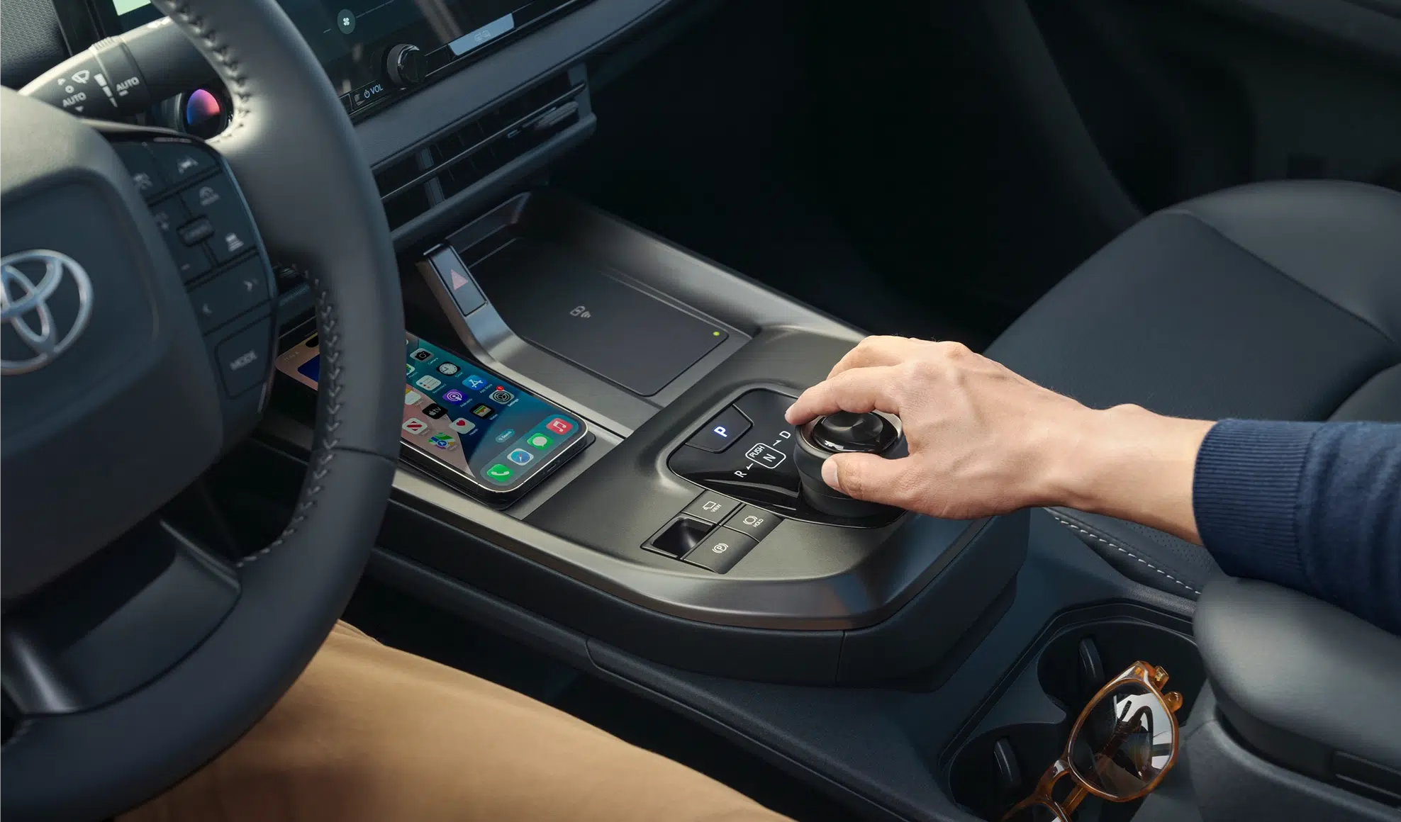 Driver's hand on the gear selector in a modern car, with a smartphone resting on the center console beside sunglasses.
