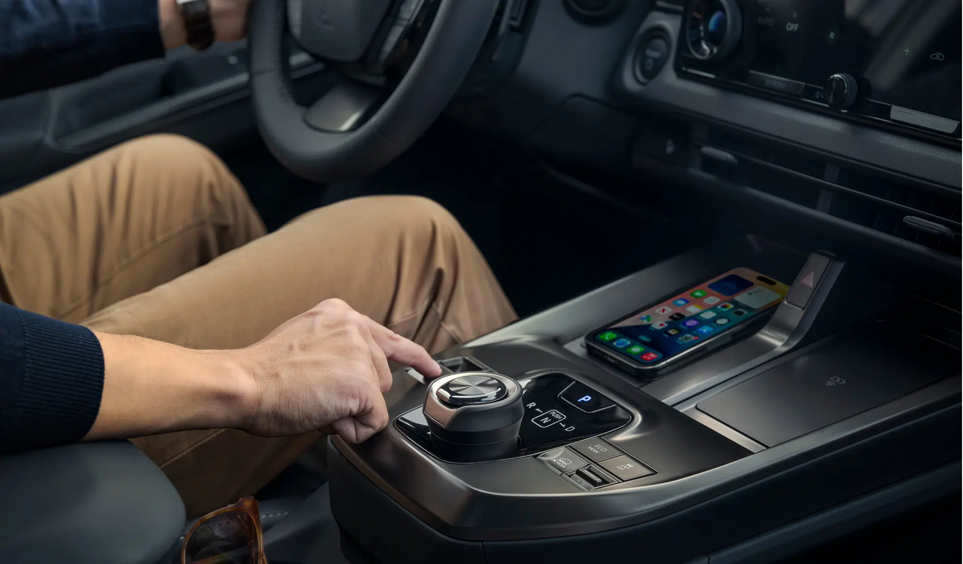 Driver’s hand on the gear selector in a car’s center console, with a smartphone resting on the console nearby behind the dash.