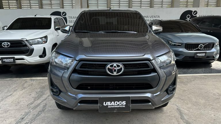 Front view of a gray Toyota pickup at a dealership, with a USADO'S Autotropical license plate and white/blue SUVs nearby.