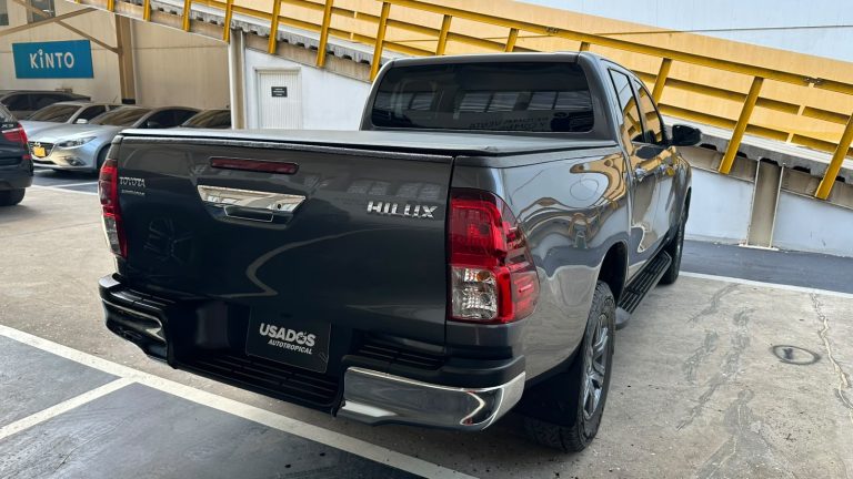 Rear three-quarter view of a dark gray Toyota Hilux pickup with a tonneau cover in a parking garage, chrome bumper, and a 'USADOS AUTOTROPICAL' plate.