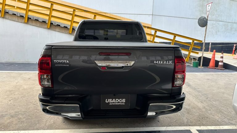 Rear view of a dark gray Toyota Hilux pickup in a parking lot, with a yellow railing and orange cones in the background.