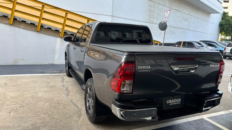 Rear view of a dark gray Toyota Hilux pickup with a tonneau cover, parked in a lot beside a white building with a yellow railing nearby.