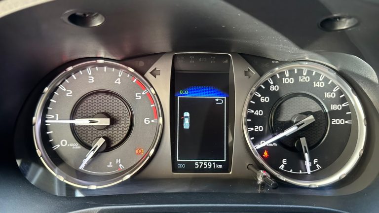 Car dashboard with analog speedometer up to 200 km/h on the right and tachometer up to 6,000 rpm on the left; center digital display shows ECO mode and a car icon, with odometer reading 57,591 km and warning indicators nearby.