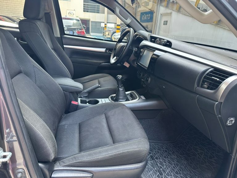 Right-hand-drive car interior with manual gear shifter, black fabric seats, center console, and touchscreen display visible on the dashboard
