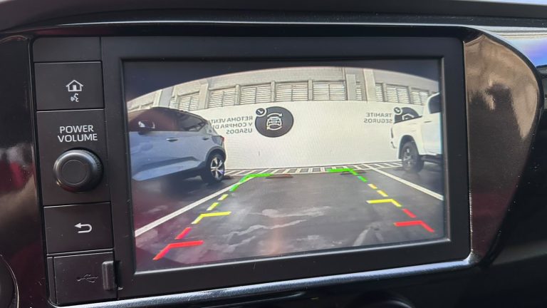 Rearview camera display showing a parking lot, with green, yellow, and red guide lines and two parked vehicles on either side near a building wall.