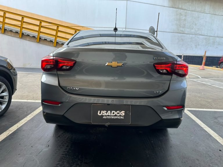 Rear view of a gray Chevrolet Onix LTZ Turbo sedan parked in a lot, showing the Chevrolet bowtie emblem on the trunk and taillights glowing red likely in daylight.