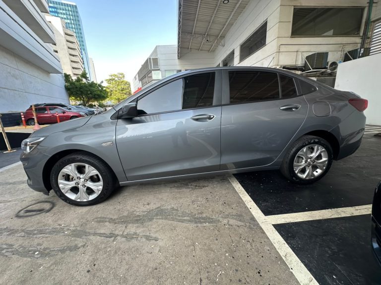 Side view of a gray four-door sedan parked in a paved parking lot beside a modern white building, with tinted windows and alloy wheels visible.