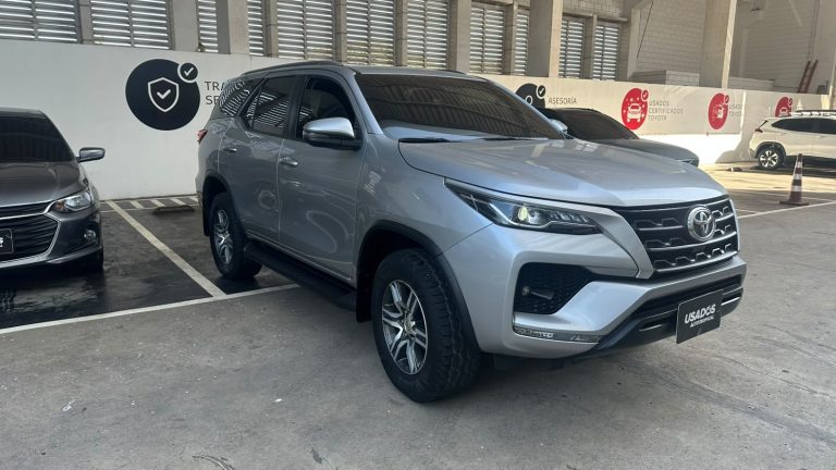 Front-right view of a silver Toyota SUV parked in a dealership showroom, with other cars nearby under a covered area.