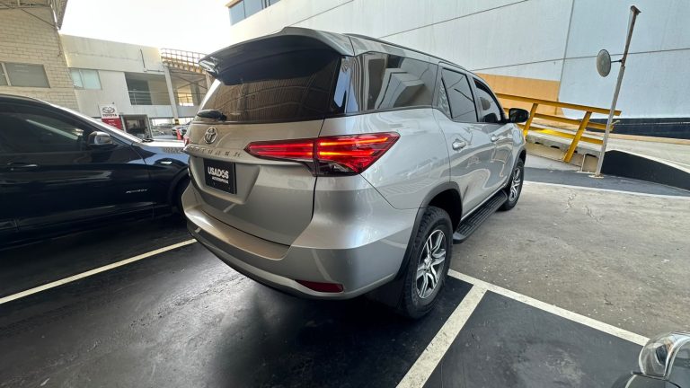 Silver Toyota SUV parked in a lot beside a dark sedan, viewed from the rear-left side with the tailgate and spoiler visible.