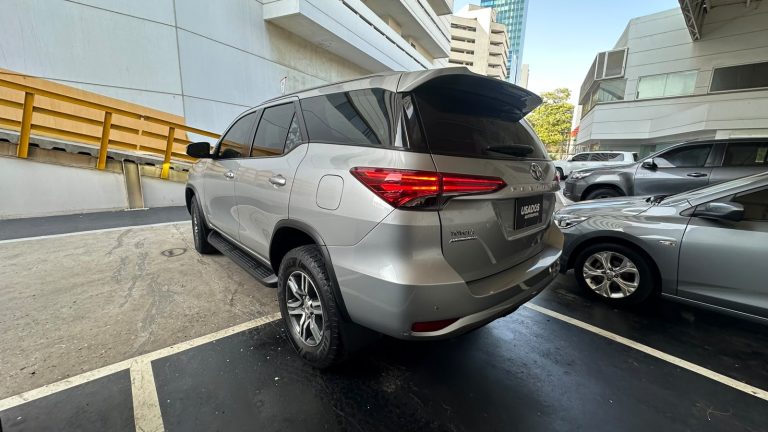 Rear three-quarter view of a silver Toyota Fortuner SUV parked in an urban parking lot beside another gray car, with modern buildings in the background and yellow railing to the left.