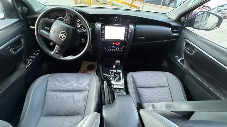 Interior of a modern sedan cabin seen from the back seat, featuring a black dashboard, steering wheel with a Toyota logo, central touchscreen, gear shifter, and gray seats with stitching.