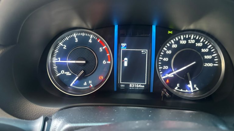 Car dashboard cluster: left tachometer, right speedometer, central screen shows parked gear (P) and 83,164 km odometer with a car icon. Blue vertical trim accents nearby.