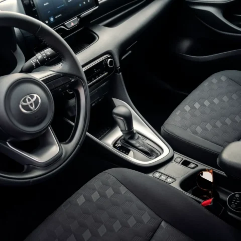 Interior of a modern car: steering wheel with Toyota logo, center console, gear shifter, and a touchscreen infotainment display visible on the dash.