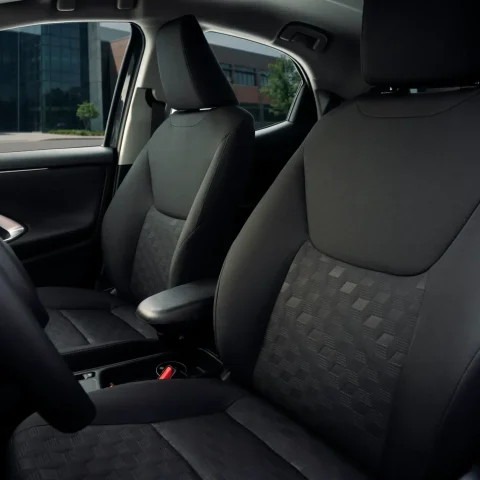Interior of a car showing two front seats with black fabric upholstery and a view of a modern building through the windows, steering wheel on the left edge.